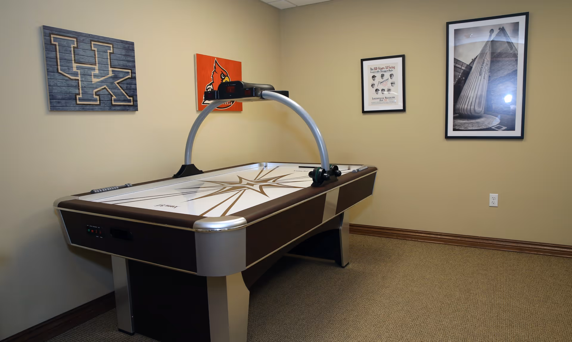 A game room with an air hockey table in the center. The walls are beige and decorated with framed pictures, including a University of Kentucky logo, a Louisville Cardinals logo, and two other framed artworks. The floor is carpeted in a neutral tone.