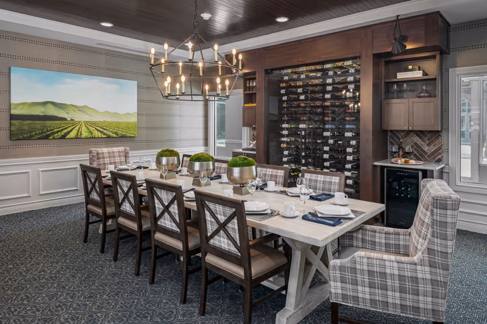 A dining room with a long wooden table set for eight people, featuring plaid upholstered chairs and three decorative green plants in silver pots as a centerpiece. The room has a patterned carpet, a large painting of a vineyard on the wall, a modern chandelier hanging from a dark wood ceiling, and a built-in wine rack with bottles displayed behind glass. There is also a small counter area with a wine fridge and shelves holding wine glasses.