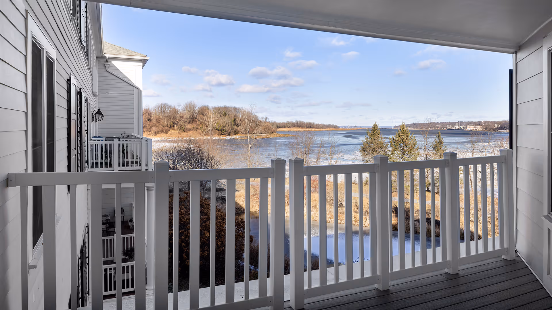 View from a balcony with white railing overlooking a partially frozen body of water, trees, and distant land under a blue sky with scattered clouds.