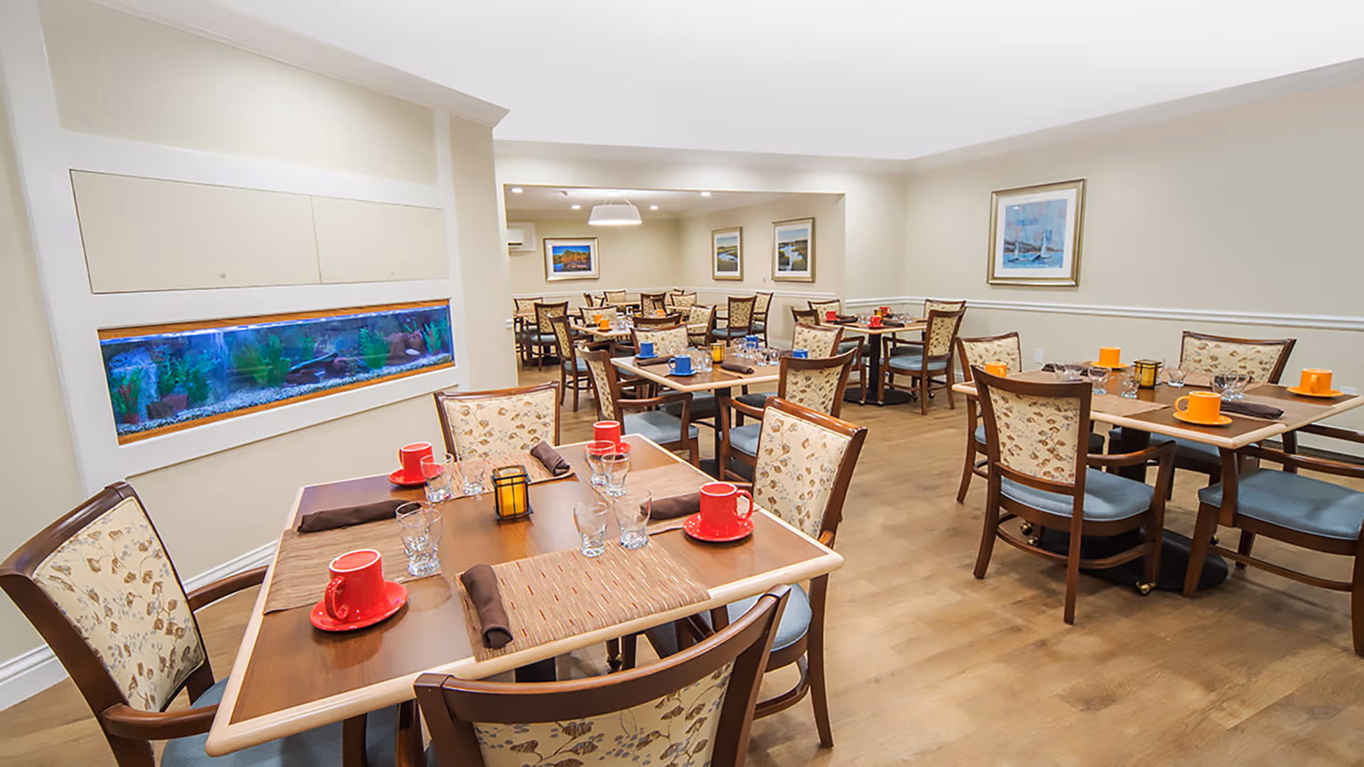 A bright and clean dining room in a senior living facility with multiple wooden tables and chairs. Each table is set with colorful cups, glasses, and napkins. There is a built-in aquarium on one wall and framed artwork hanging on the walls. The floor is wooden, and the room has a warm, inviting atmosphere.