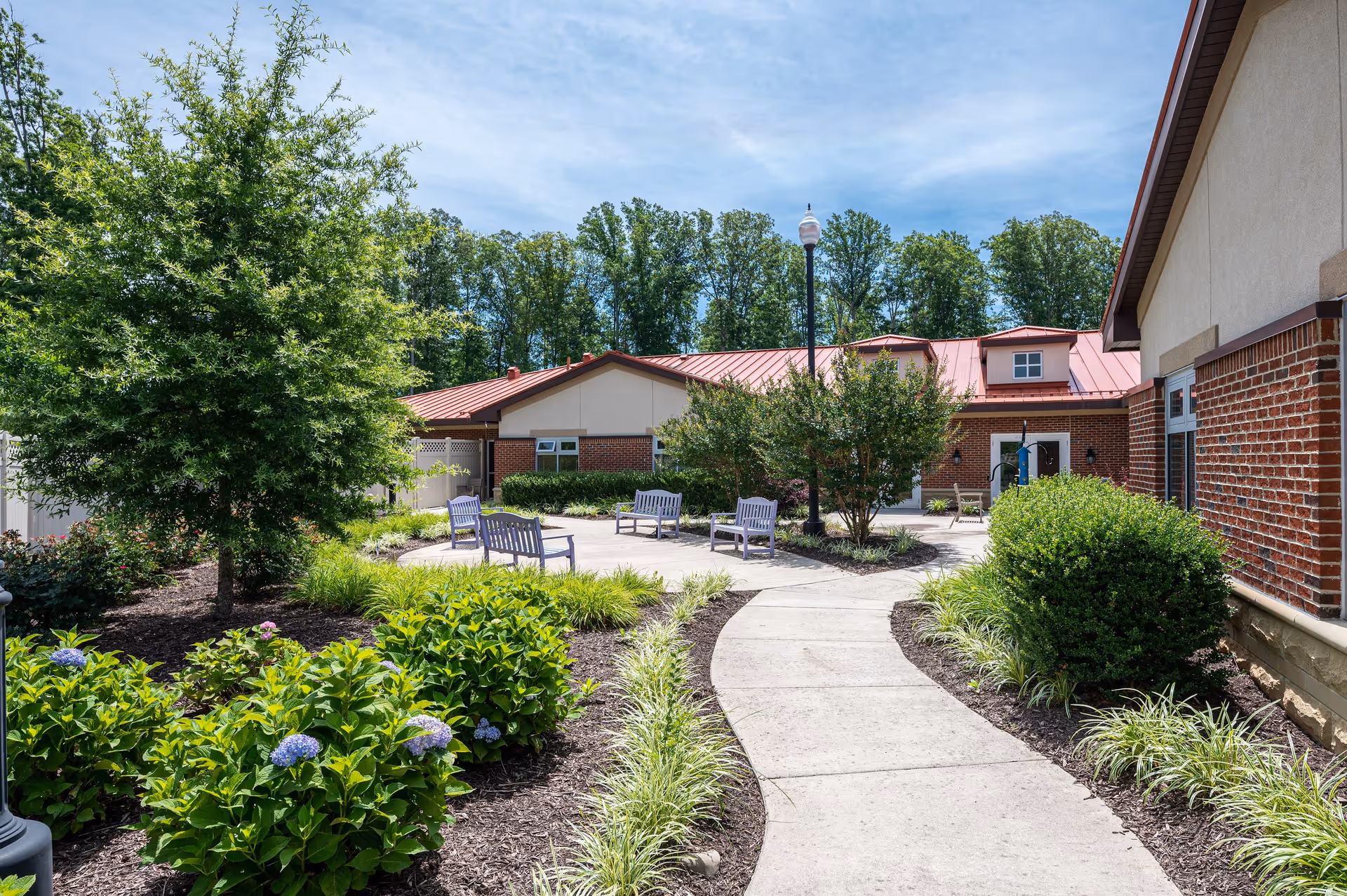 Outdoor courtyard area at Arden Courts Richmond featuring a curved concrete walkway surrounded by landscaped greenery, bushes, and flowering plants. Several benches are placed around the courtyard near a lamppost, with a brick building and trees in the background under a partly cloudy sky.