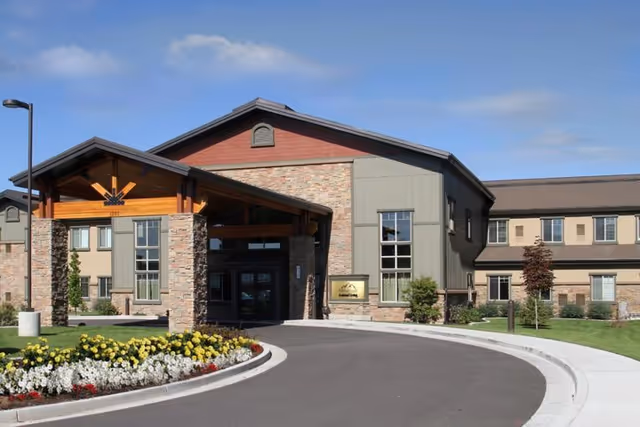 Front exterior view of Fairfield Village a Generations Community building with a covered entrance, stone and wood facade, landscaped flower beds, and a curved driveway under a partly cloudy sky.