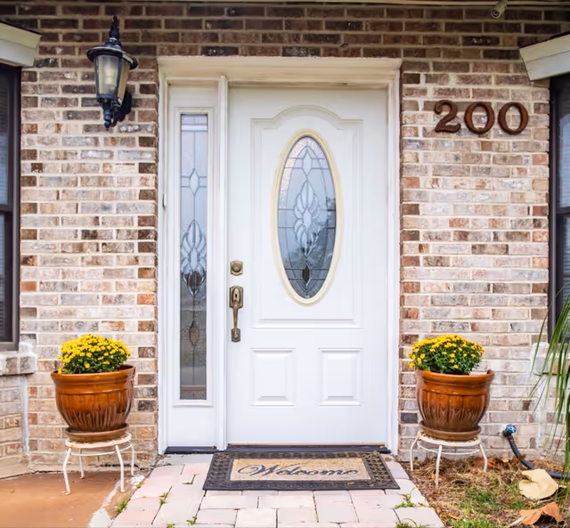 Front entrance of a brick building with a white door featuring decorative oval glass panels. Two brown flower pots with yellow flowers are placed on white stands on either side of the door. A black outdoor lantern is mounted on the left side of the door. The number 200 is displayed on the right side of the door. A welcome mat is placed in front of the door on a paved walkway.