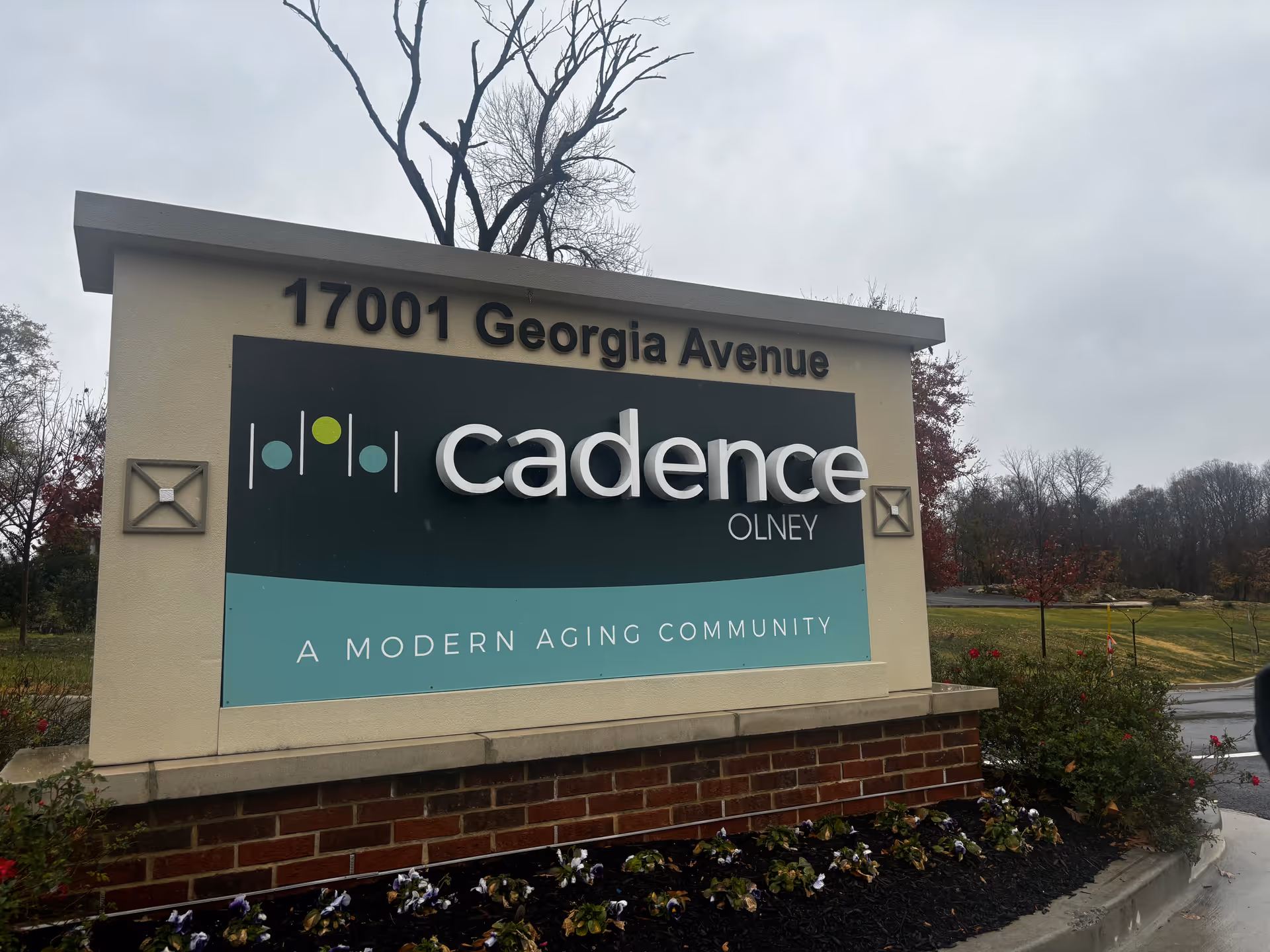 Outdoor sign for Cadence Olney, a modern aging community, located at 17001 Georgia Avenue. The sign is set in a landscaped area with flowers and bushes, with a cloudy sky and trees in the background.