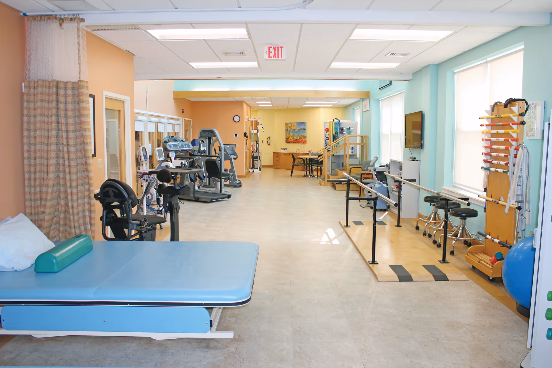 Bright rehabilitation/physical therapy room with exercise equipment, parallel bars, a treatment table and seating.