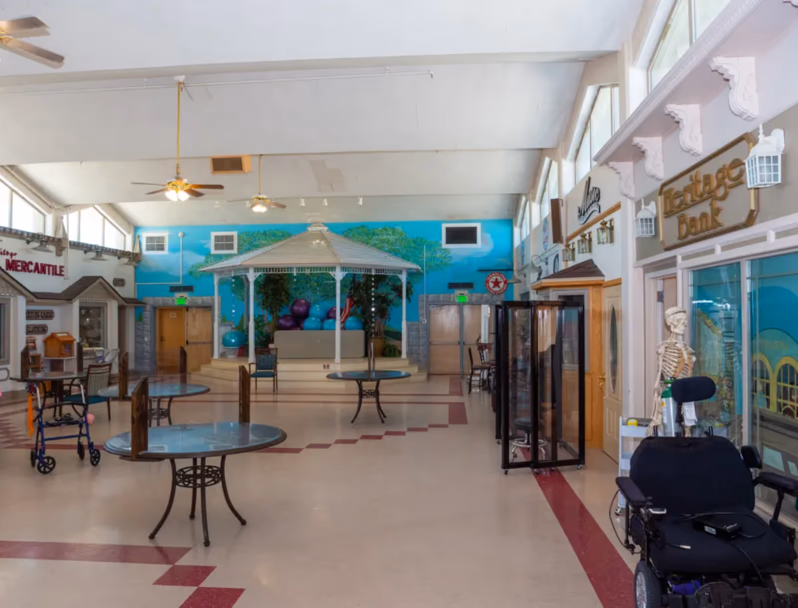Interior view of a spacious common area in a senior living facility with round tables, chairs, a gazebo structure in the center, and storefront-style setups labeled Heritage Mercantile and Heritage Bank. There is a skeleton model and a motorized wheelchair visible on the right side. The walls are decorated with murals and the ceiling has ceiling fans and lights.