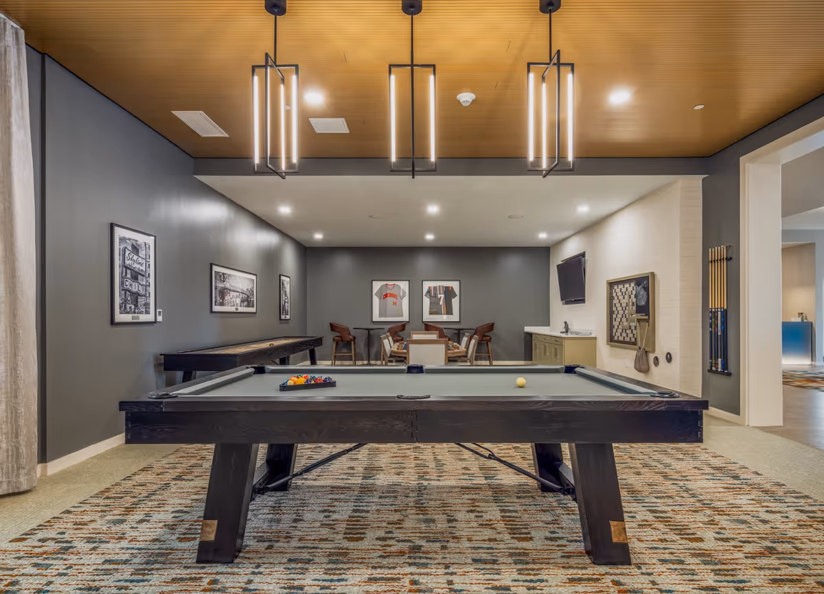 A recreational room featuring a pool table in the foreground with billiard balls arranged on it. Behind the pool table, there is a shuffleboard table along the left wall and a seating area with chairs and a table. The walls are decorated with framed pictures and sports jerseys. On the right wall, there is a mounted TV above a small counter with a sink, and a wall-mounted chessboard with a phone below it. The room has a patterned carpet, modern pendant lights hanging from a wooden ceiling, and a neutral color scheme.