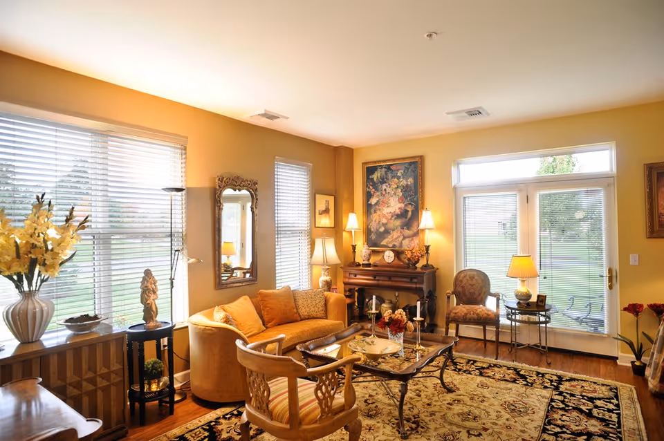 Sunlit traditional living room with a sofa, armchairs, coffee table, lamps, artwork, and large windows/glass doors overlooking a lawn.
