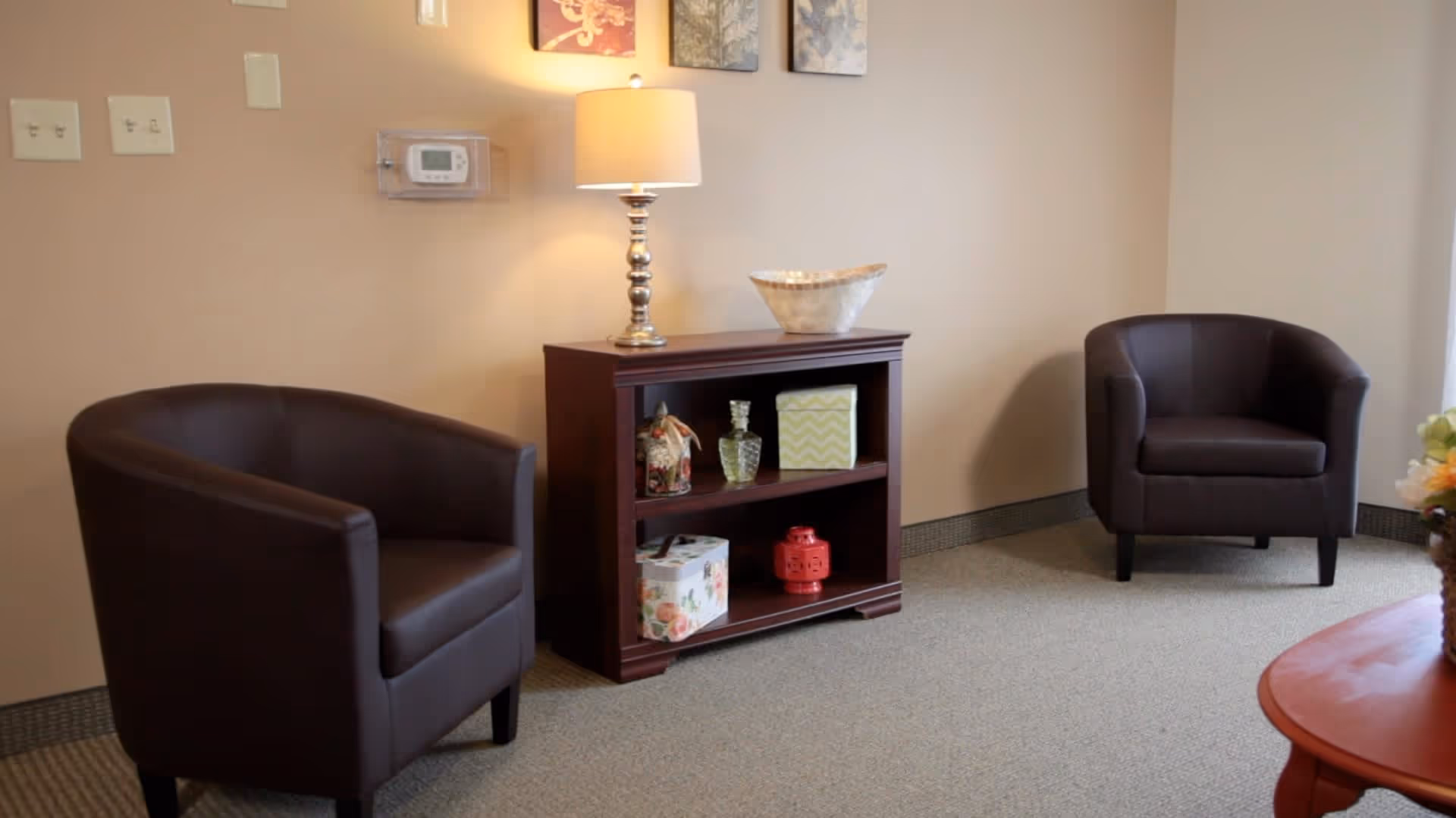 A cozy sitting area with two dark brown leather armchairs facing each other, a small wooden bookshelf between them holding decorative items, and a table lamp on top. The walls are beige, and there is a thermostat on the wall above the bookshelf.