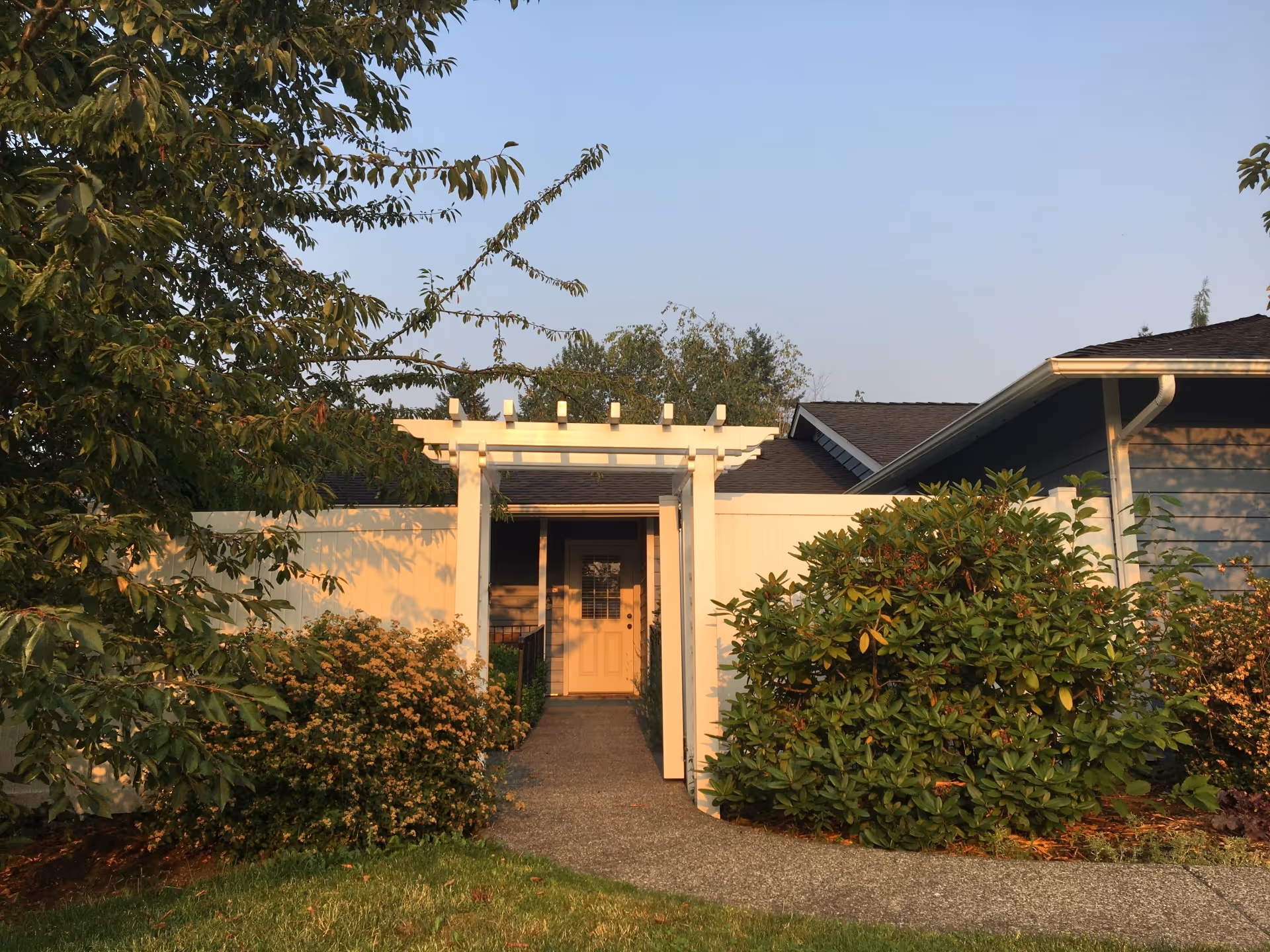 Front entrance of a single-story building with a white pergola over a walkway, flanked by shrubs and trees.