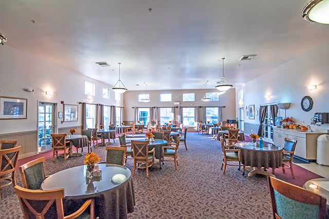 A spacious dining room with multiple round tables covered with dark tablecloths and surrounded by wooden chairs. Each table has a small floral centerpiece. The room has large windows letting in natural light, light fixtures hanging from the ceiling, and framed pictures on the walls. There is a clock and a beverage station on one side of the room.