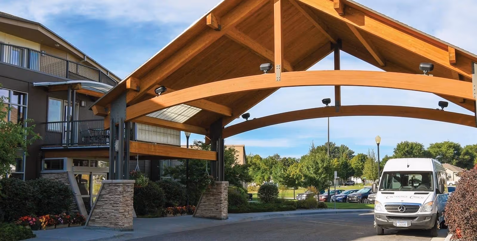 Entrance area of St. John's United - The Crossings facility featuring a large wooden canopy supported by stone pillars, with a white Mercedes van parked nearby and a multi-story building in the background surrounded by greenery and a clear sky.