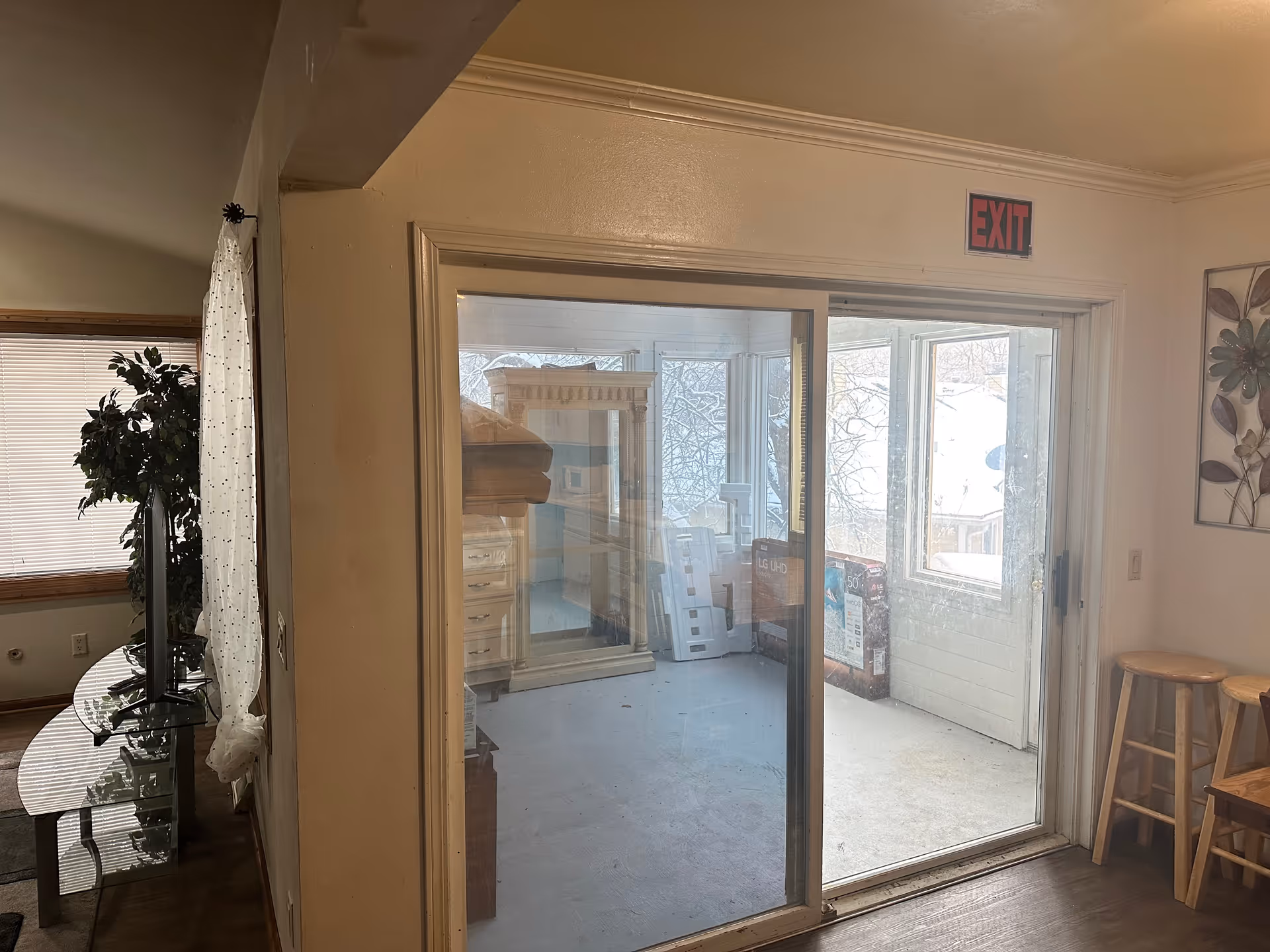 Interior view showing a sliding glass door leading to a sunroom or enclosed porch with some furniture and boxes inside. To the right of the door are two wooden stools and a wall decoration with floral design. To the left, part of a living room area is visible with a TV on a glass stand and a large potted plant near a window with blinds and a sheer curtain.