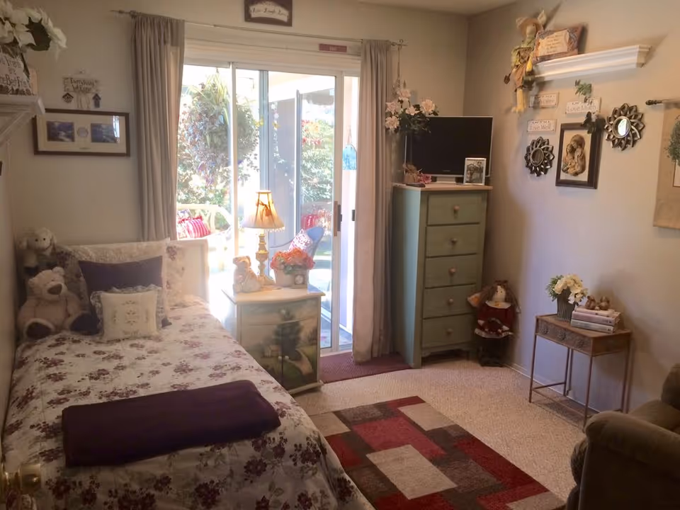 A cozy bedroom with a single bed covered in floral bedding and pillows, a nightstand with a lamp and decorative items, a green chest of drawers with a small TV on top, and various wall decorations including framed pictures and mirrors. There is a sliding glass door leading to an outdoor area with plants visible outside.