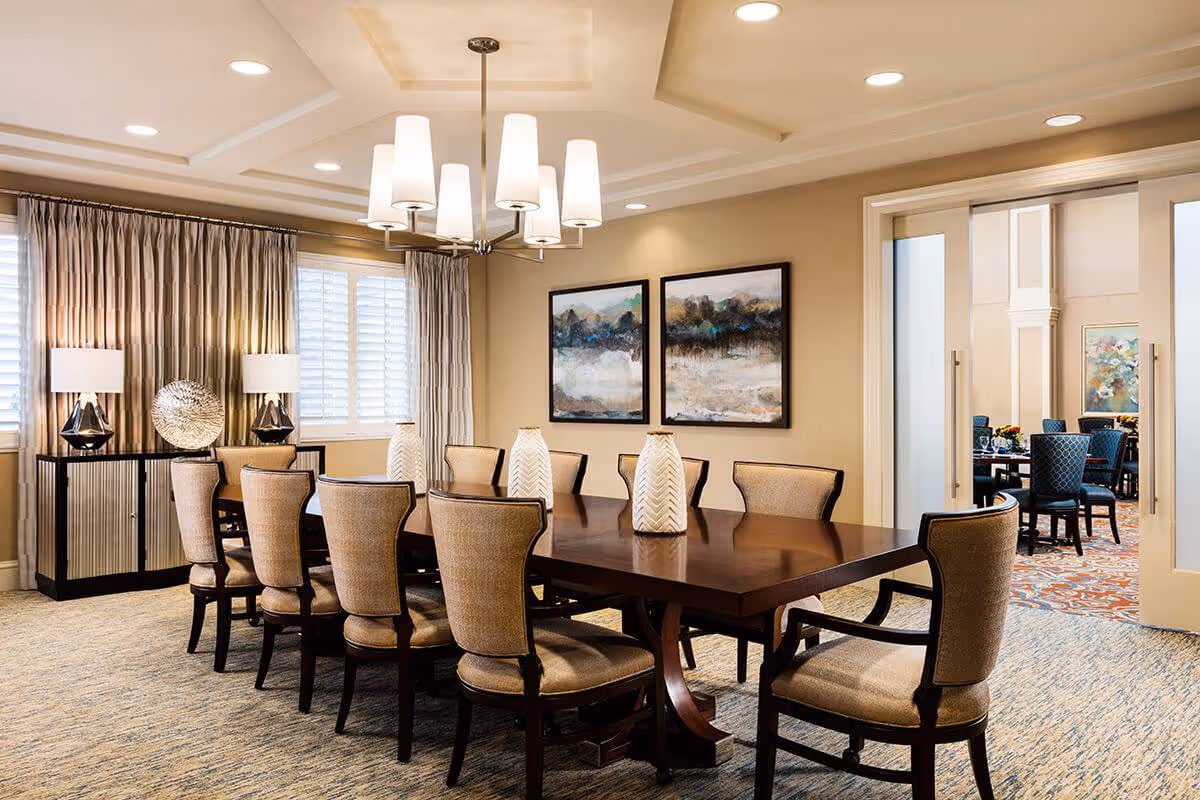 A well-lit dining room with a large wooden table surrounded by ten upholstered chairs. The room features a modern chandelier hanging from a coffered ceiling, two abstract paintings on the beige wall, and a sideboard with decorative lamps and a metallic sculpture. Adjacent to this room is another dining area visible through sliding glass doors.