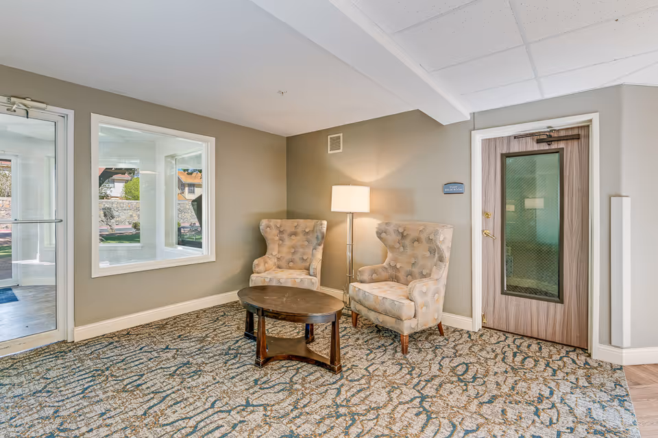 A small seating area in a senior living facility with two patterned armchairs, a round wooden coffee table, and a floor lamp. The walls are painted light gray, and there is a door labeled 'Staff Break Room' on the right side. A window and glass door on the left show an outdoor view.
