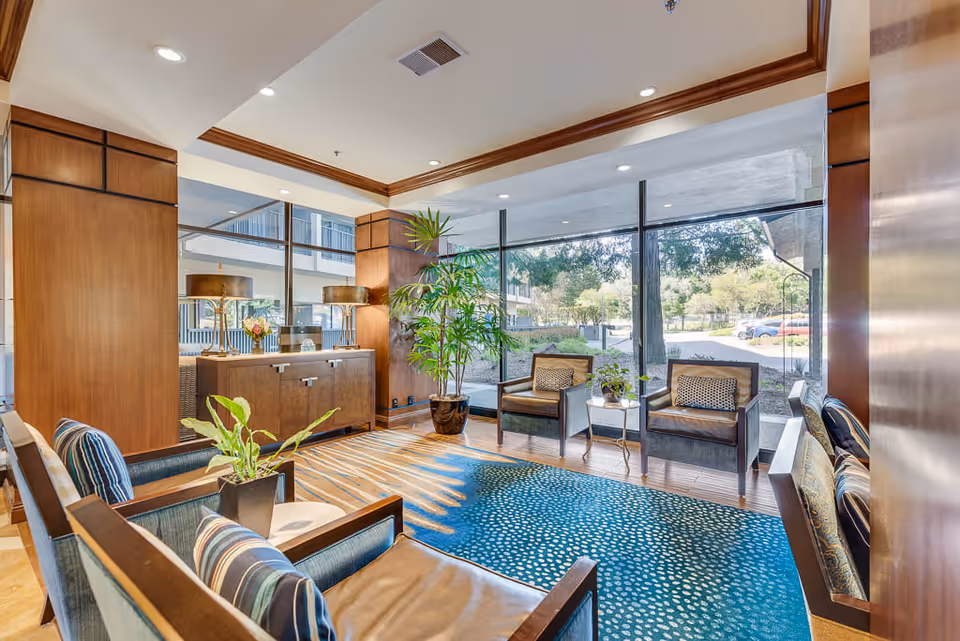 A bright and inviting senior living facility lounge area with large floor-to-ceiling windows showing outdoor greenery. The room features wooden paneling, a blue patterned rug, several armchairs with cushions, a wooden sideboard with two lamps, and potted plants.