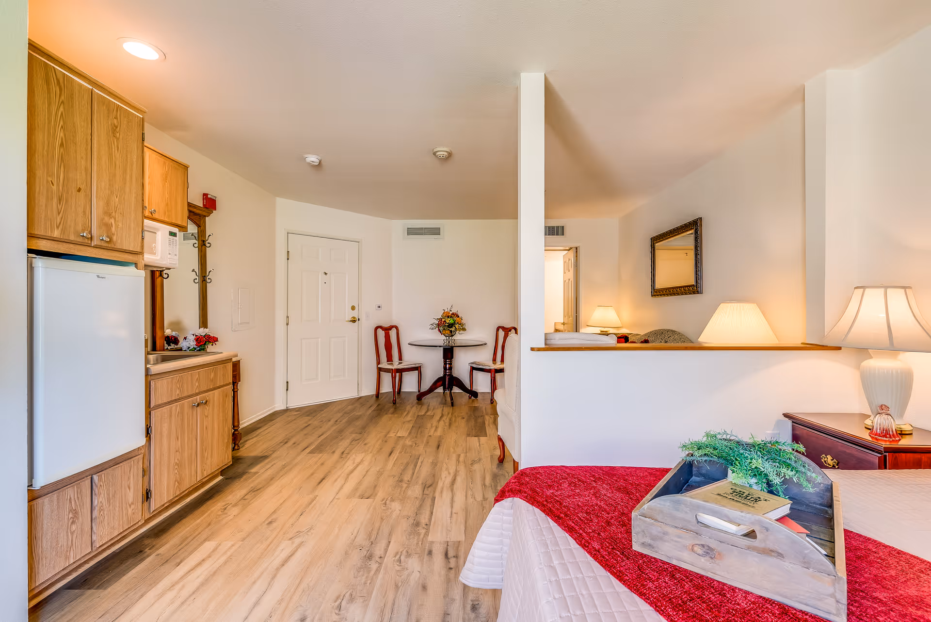 Interior view of a senior assisted living unit featuring a small kitchenette with wooden cabinets, a white refrigerator, and a microwave. There is a round table with two chairs near the entrance door. The room has wood flooring and a bed with a red blanket and a wooden tray holding a book and a plant. A lamp and a mirror are visible on the right side of the room.