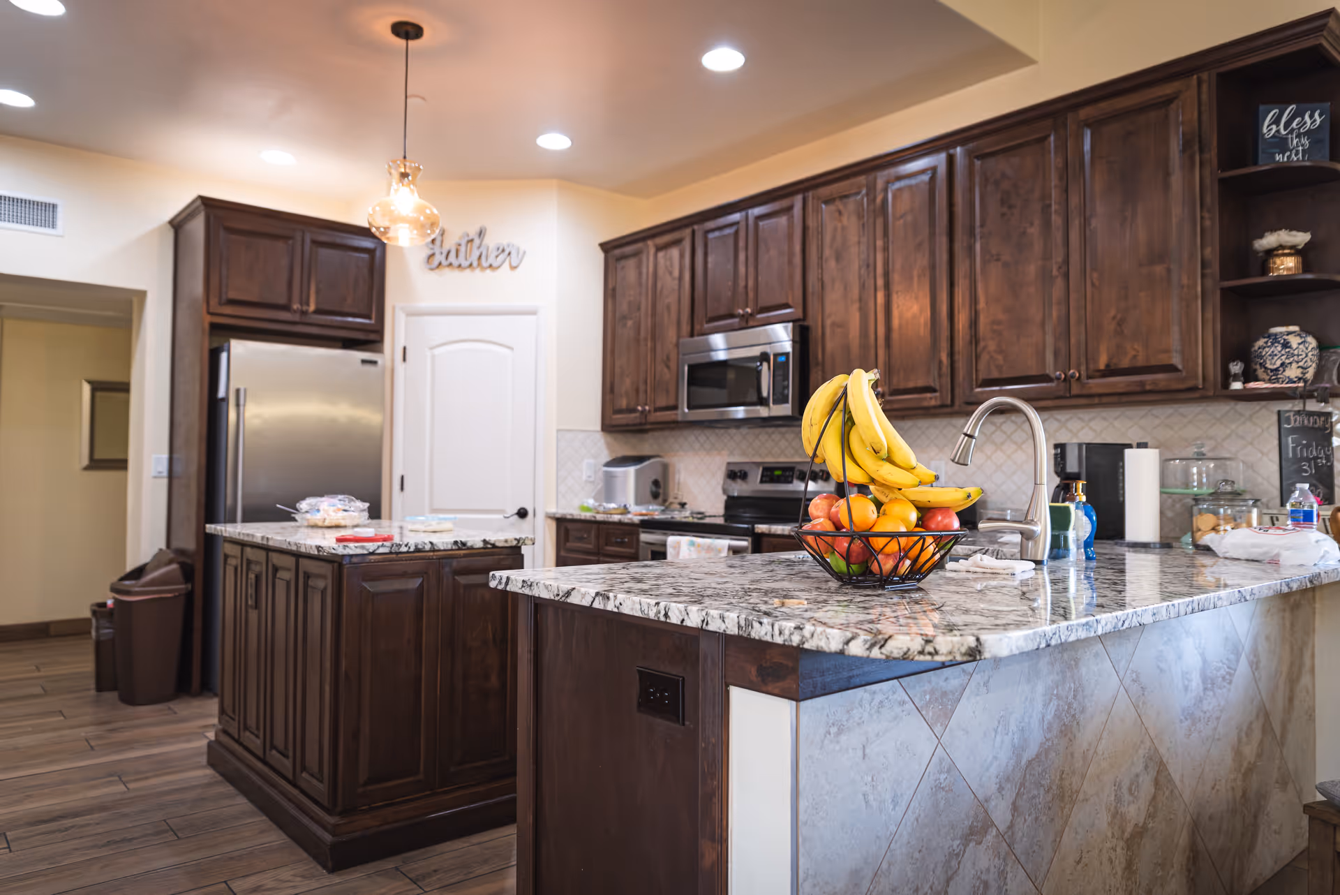 Modern kitchen with dark wooden cabinets, granite countertops, a stainless steel refrigerator, microwave, and stove. A fruit basket with bananas and apples sits on the kitchen island. The kitchen has pendant lighting and a sign on the wall that says 'Gather'.