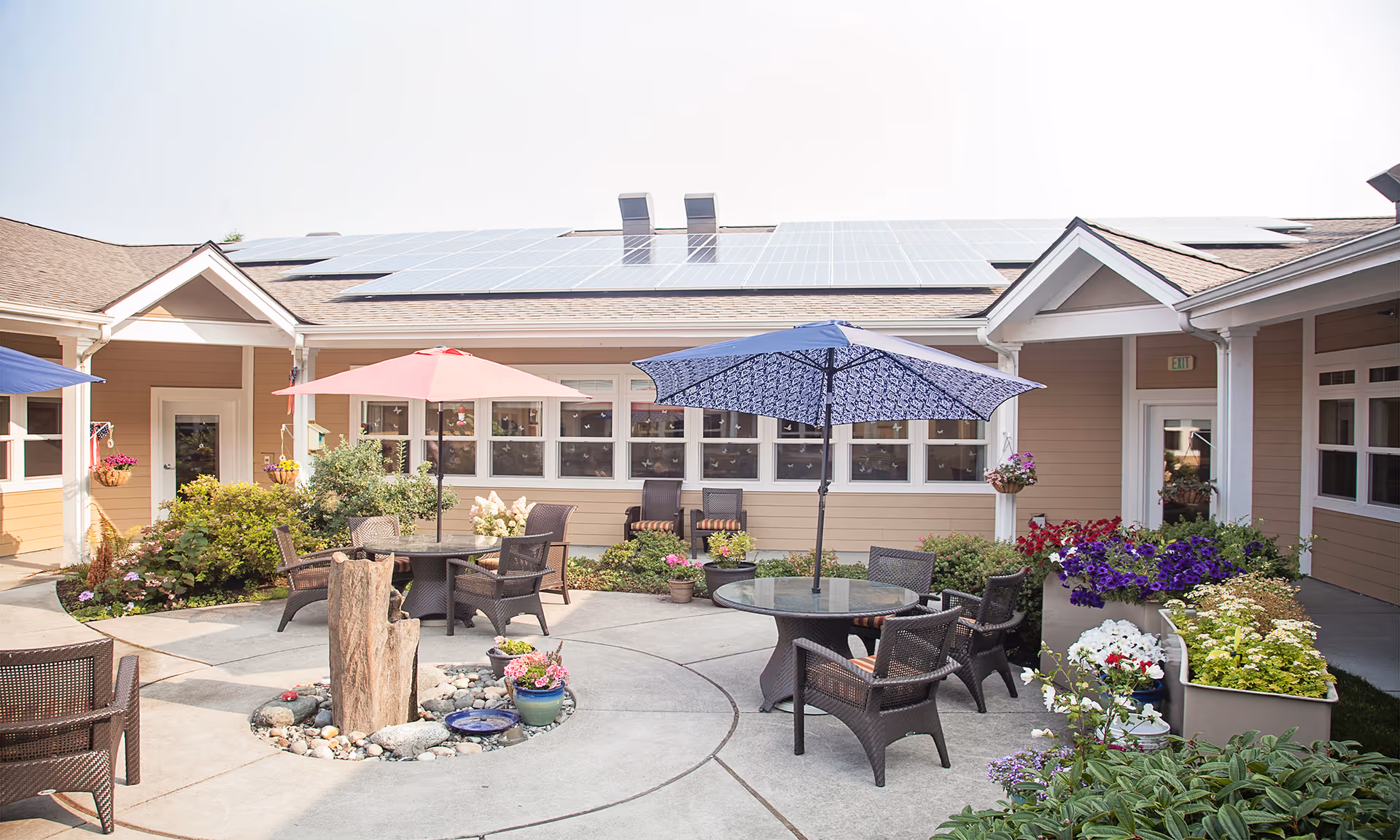Sunny courtyard with round glass-top patio tables, umbrellas and wicker chairs surrounded by flower beds in front of a single-story building with solar panels.