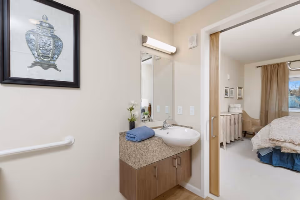 A bathroom area with a wall-mounted sink, a mirror above it, a blue towel on the countertop, and a framed artwork on the wall. To the right, there is an open sliding door leading to a bedroom with a bed, a chair, and a window with curtains.