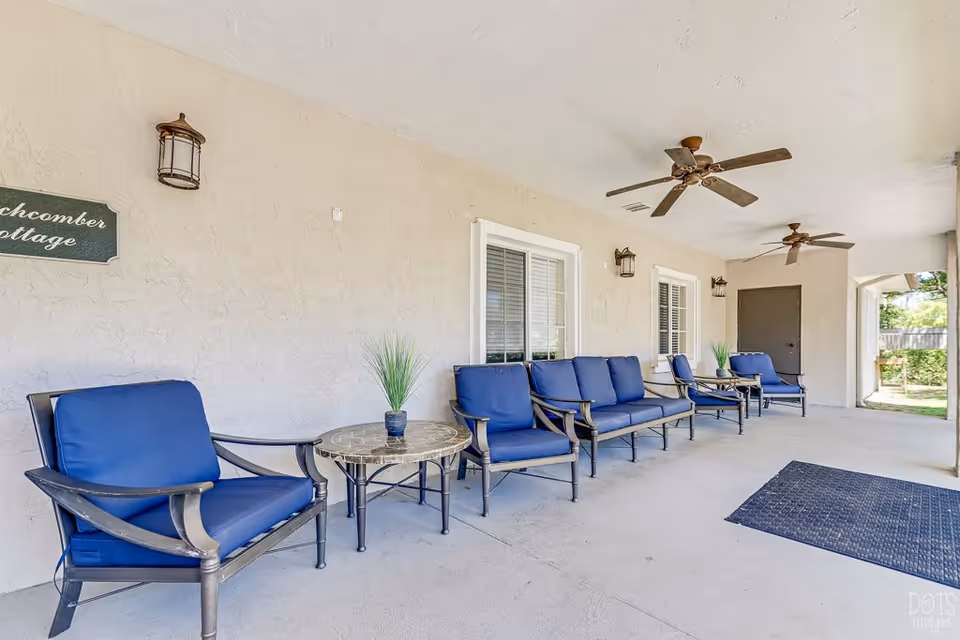 Covered outdoor patio area with blue cushioned chairs and sofas arranged around small round tables with potted plants. The patio has ceiling fans, wall-mounted lantern lights, and a beige textured wall with windows and a door. A sign on the wall reads 'Beachcomber Cottage'.