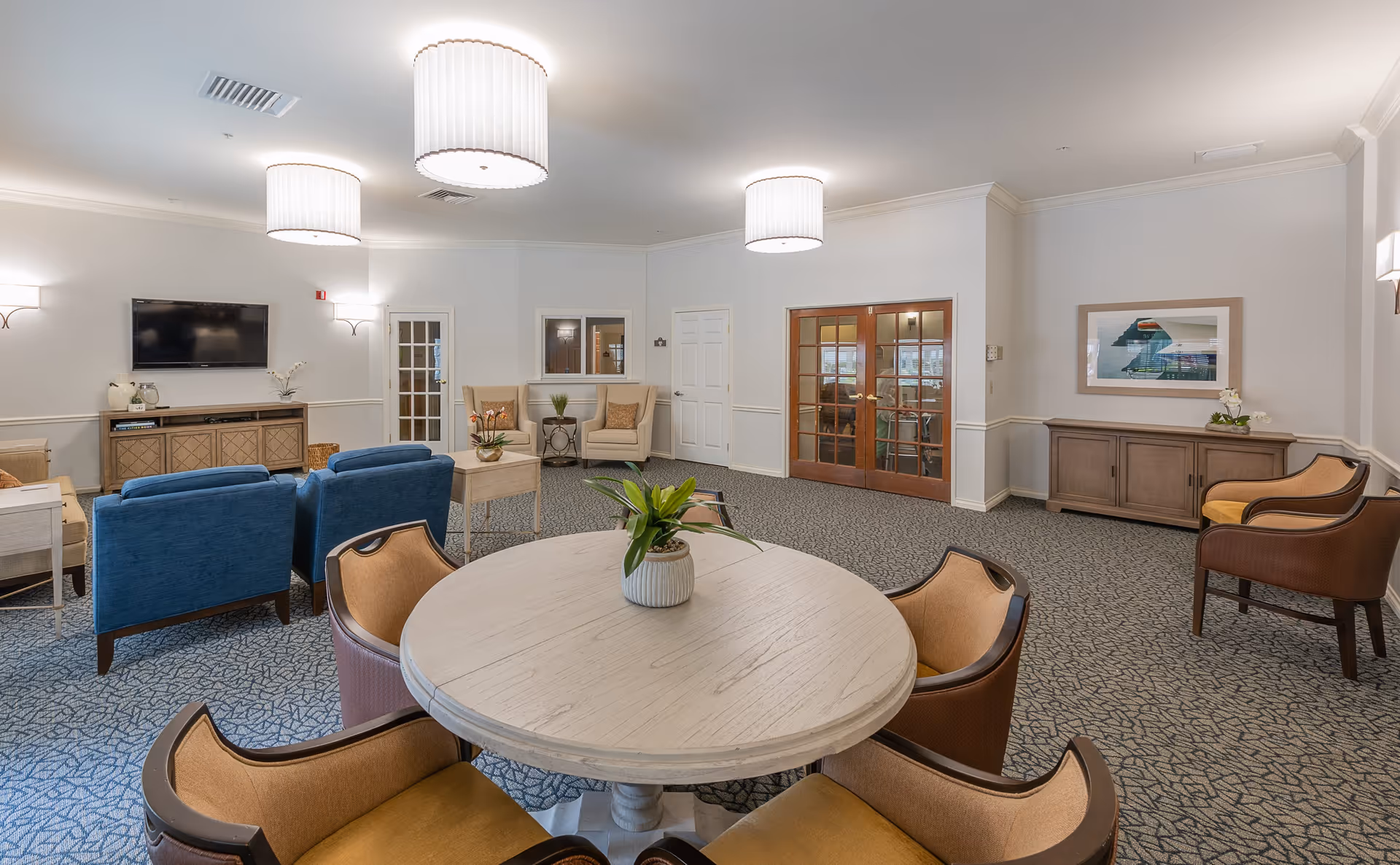 A spacious and well-lit common living area with a round table and four chairs in the foreground, blue armchairs, beige armchairs, a wall-mounted TV, wooden cabinets, and decorative plants. The room features patterned carpet, white walls, and ceiling lights.