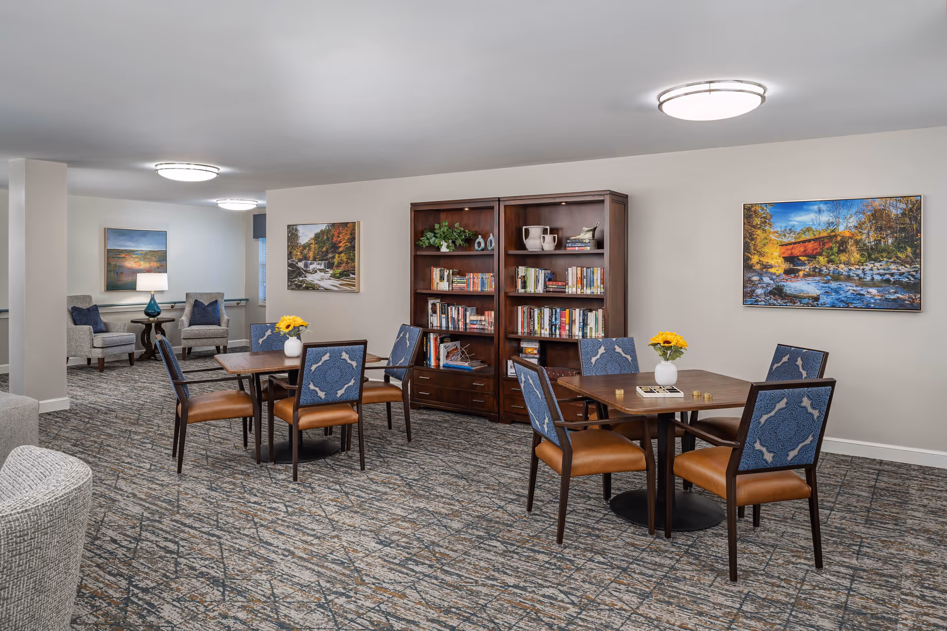 A senior living facility common area with two wooden tables, each surrounded by four chairs with blue patterned backs and brown seats. Two wooden bookshelves filled with books and decorative items are against the wall. The room has carpeted flooring with a geometric pattern, neutral-colored walls, and ceiling lights. There are framed landscape paintings on the walls and a small seating area with two gray armchairs and a side table with a lamp in the background.