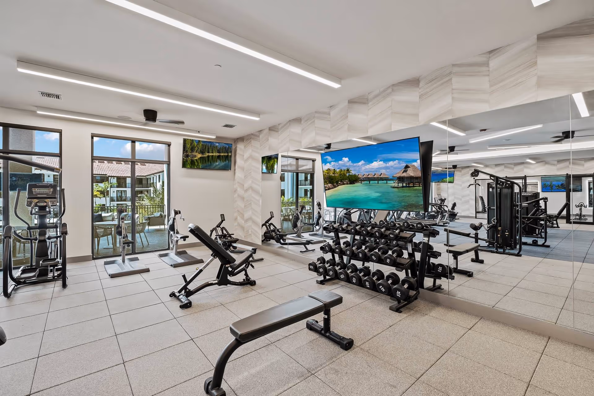 Modern fitness room with dumbbell racks, benches, cardio machines, mirrored wall and a large TV displaying a beach scene.