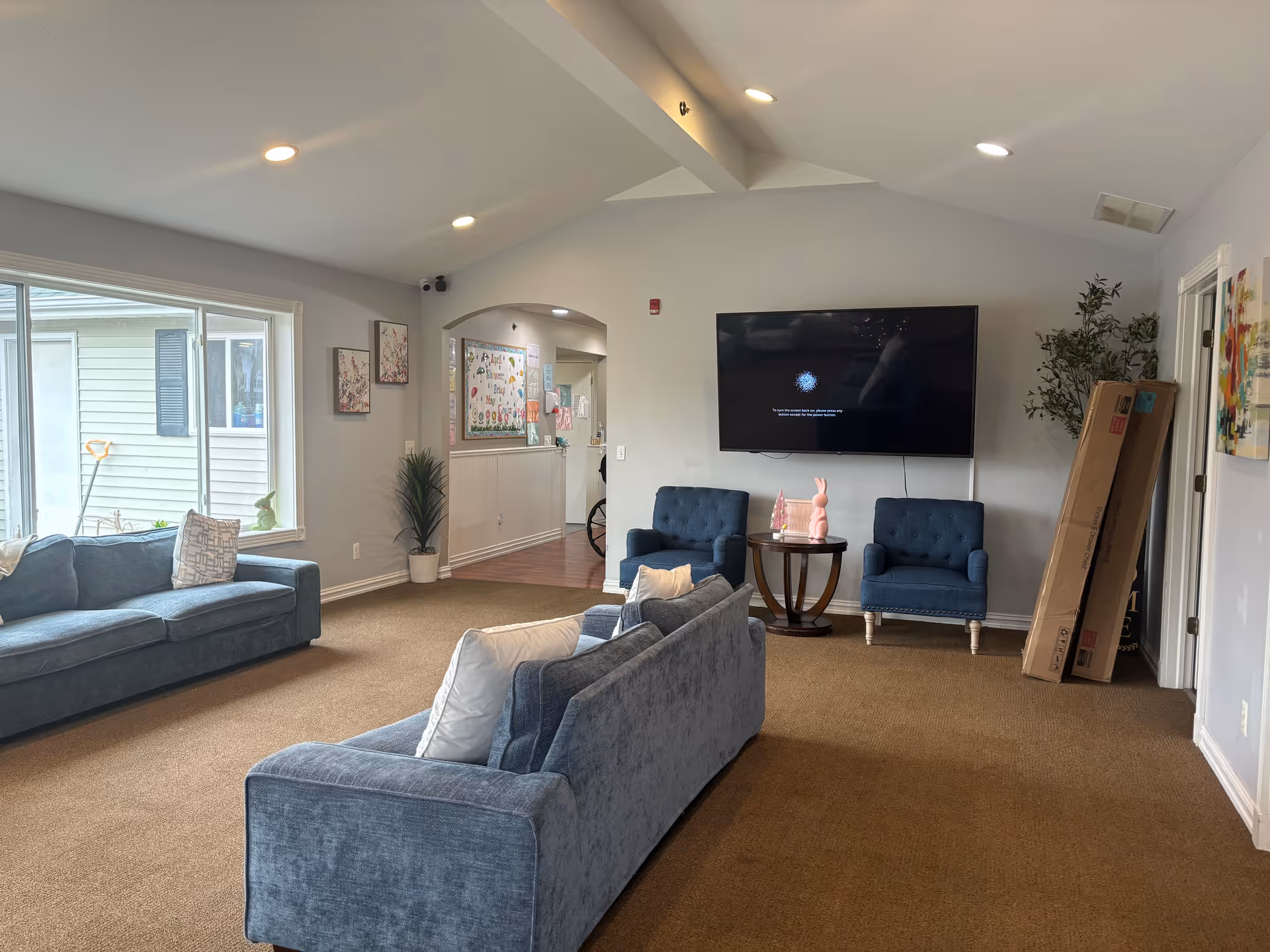 Communal living room with blue sofas and armchairs, a wall-mounted TV, coffee table, and a large window.