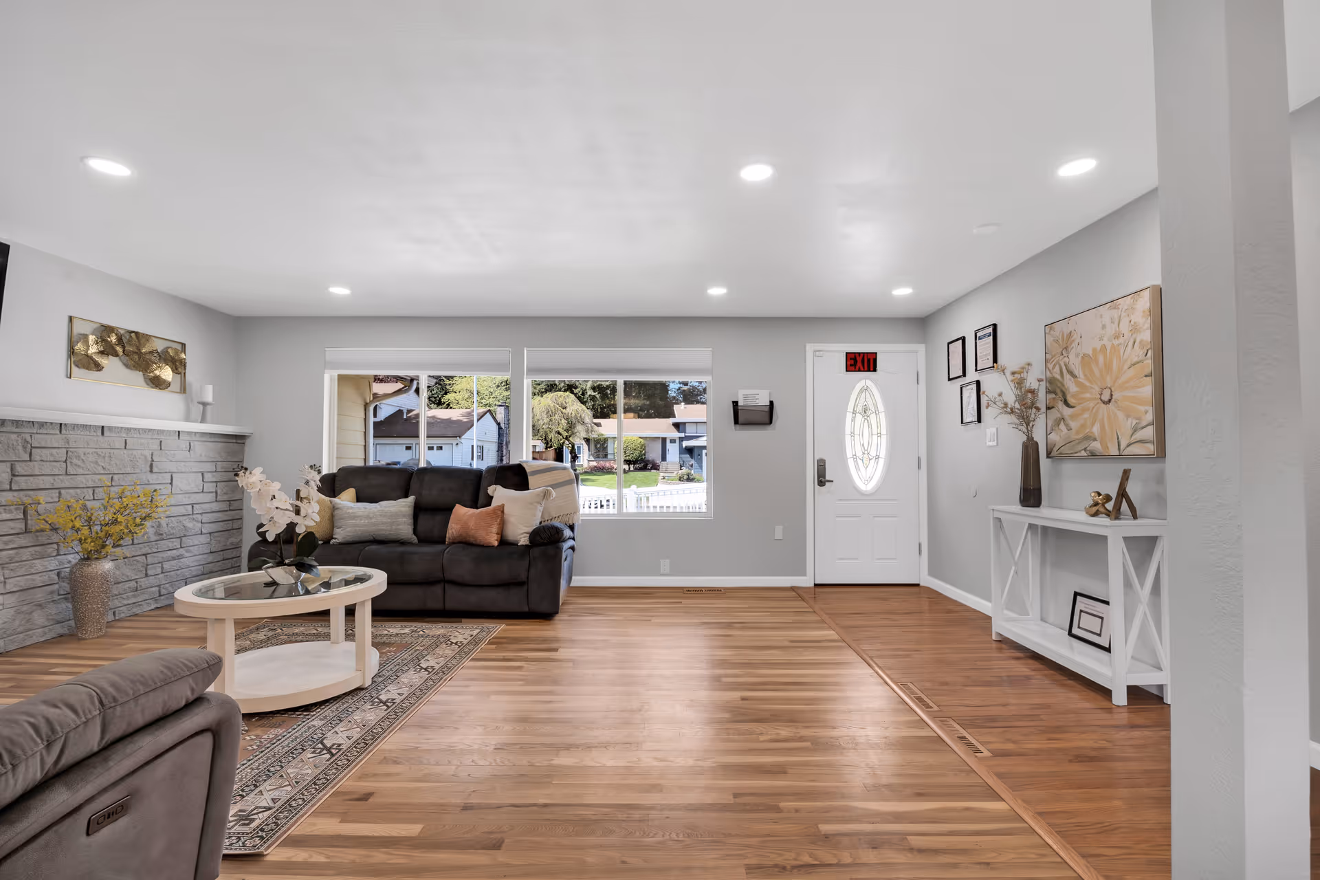 A bright living room with light gray walls and wooden flooring. The room features a dark gray sofa with multiple cushions, a round white coffee table with a glass top and decorative flowers, and a patterned rug. There is a stone fireplace on the left wall with a vase of yellow flowers on the floor. The far wall has two large windows showing a view of houses and trees outside. To the right, there is a white front door with an oval glass panel and an exit sign above it. A white console table with framed pictures and a vase with dried flowers is against the right wall. The ceiling has recessed lighting.