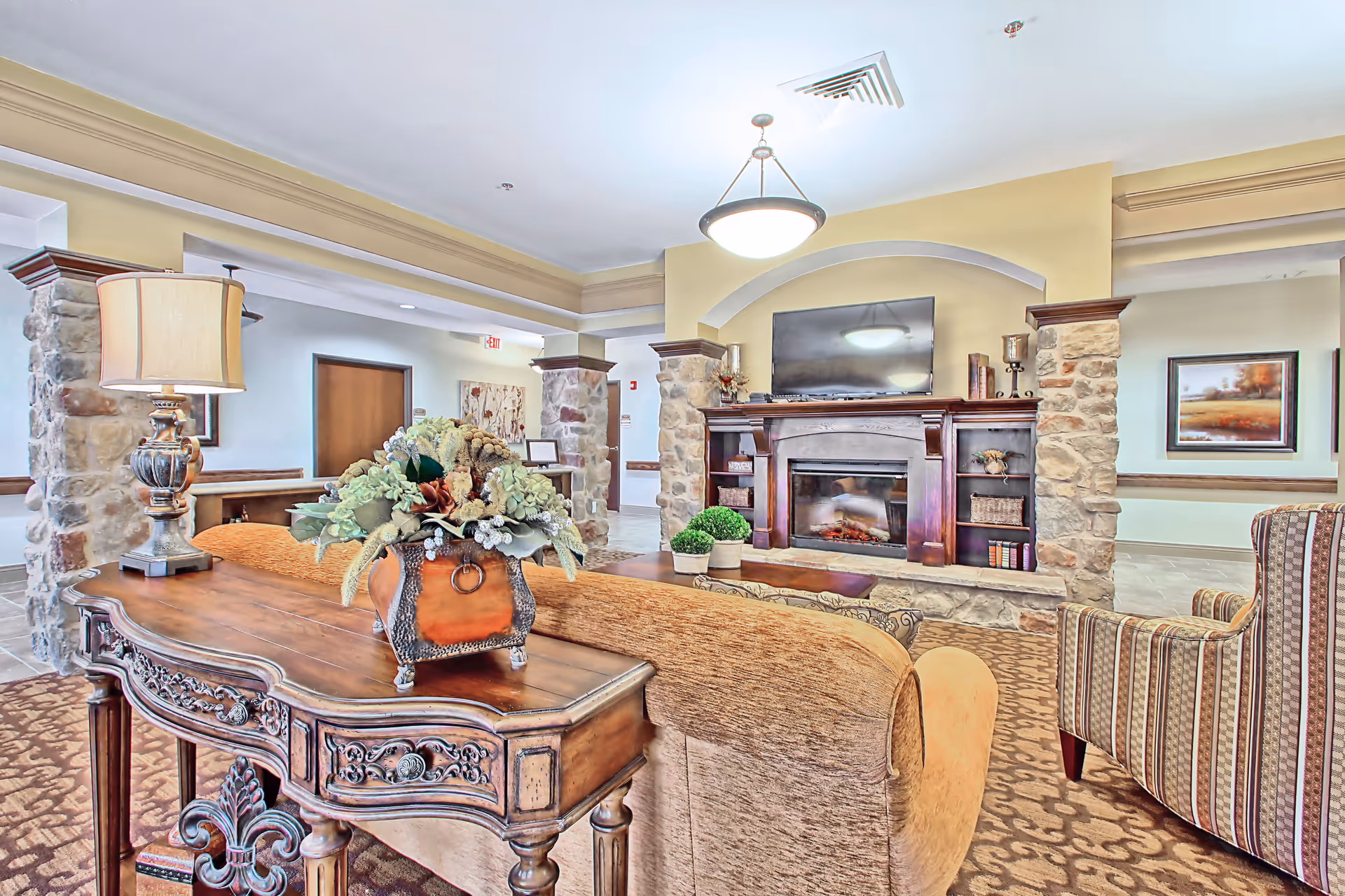 A cozy living room area in a senior living facility featuring a stone fireplace with a mounted flat-screen TV above it. The room has a wooden console table with a decorative floral arrangement and a lamp. There are comfortable upholstered chairs and a sofa arranged around the fireplace. The walls are painted light yellow and beige, with stone pillars and framed artwork visible in the background.
