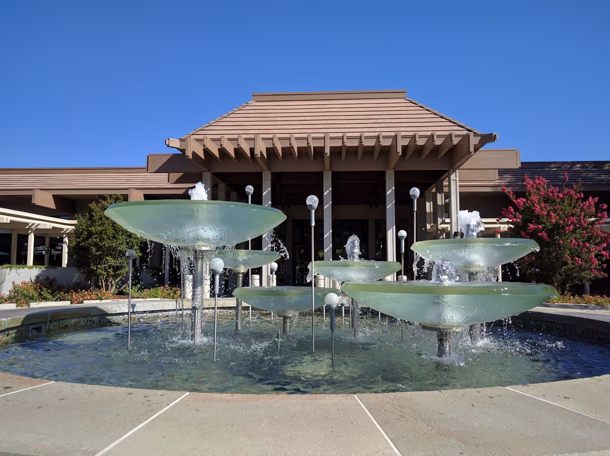 Outdoor water fountain with multiple glass bowls and water jets in front of a building with a brown tiled roof and columns, surrounded by greenery and flowering bushes under a clear blue sky.