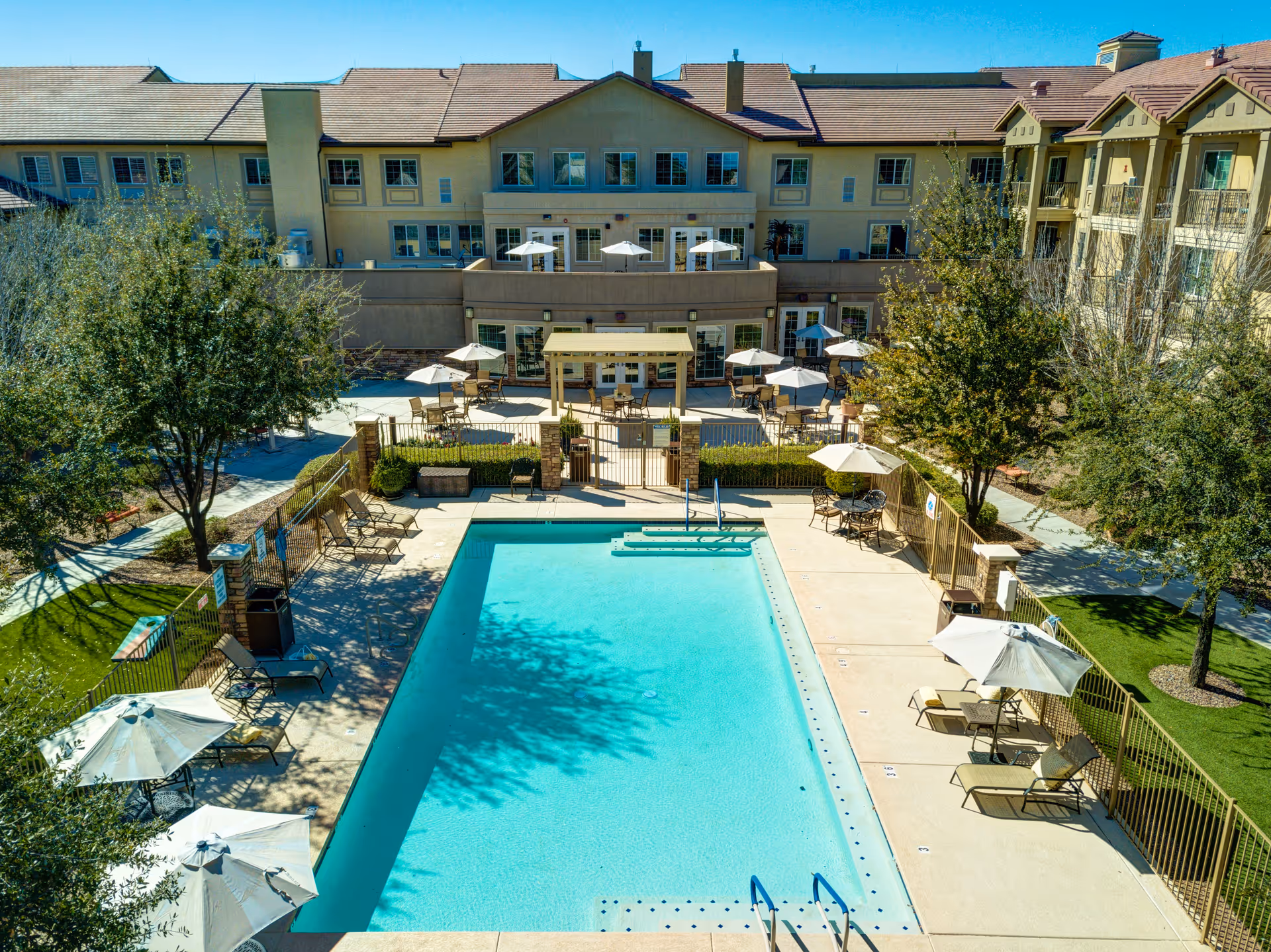 Outdoor swimming pool area at a senior living facility with lounge chairs and umbrellas surrounding the pool. Trees and green lawns are visible around the pool area, with a multi-story building in the background featuring balconies and windows.