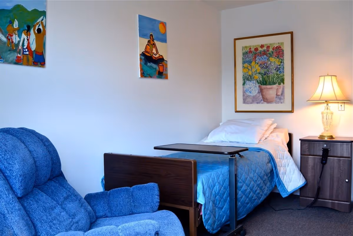 A small furnished bedroom with a single bed covered in a blue quilt, an overbed table, a blue recliner, a nightstand with a lamp, and framed wall art.