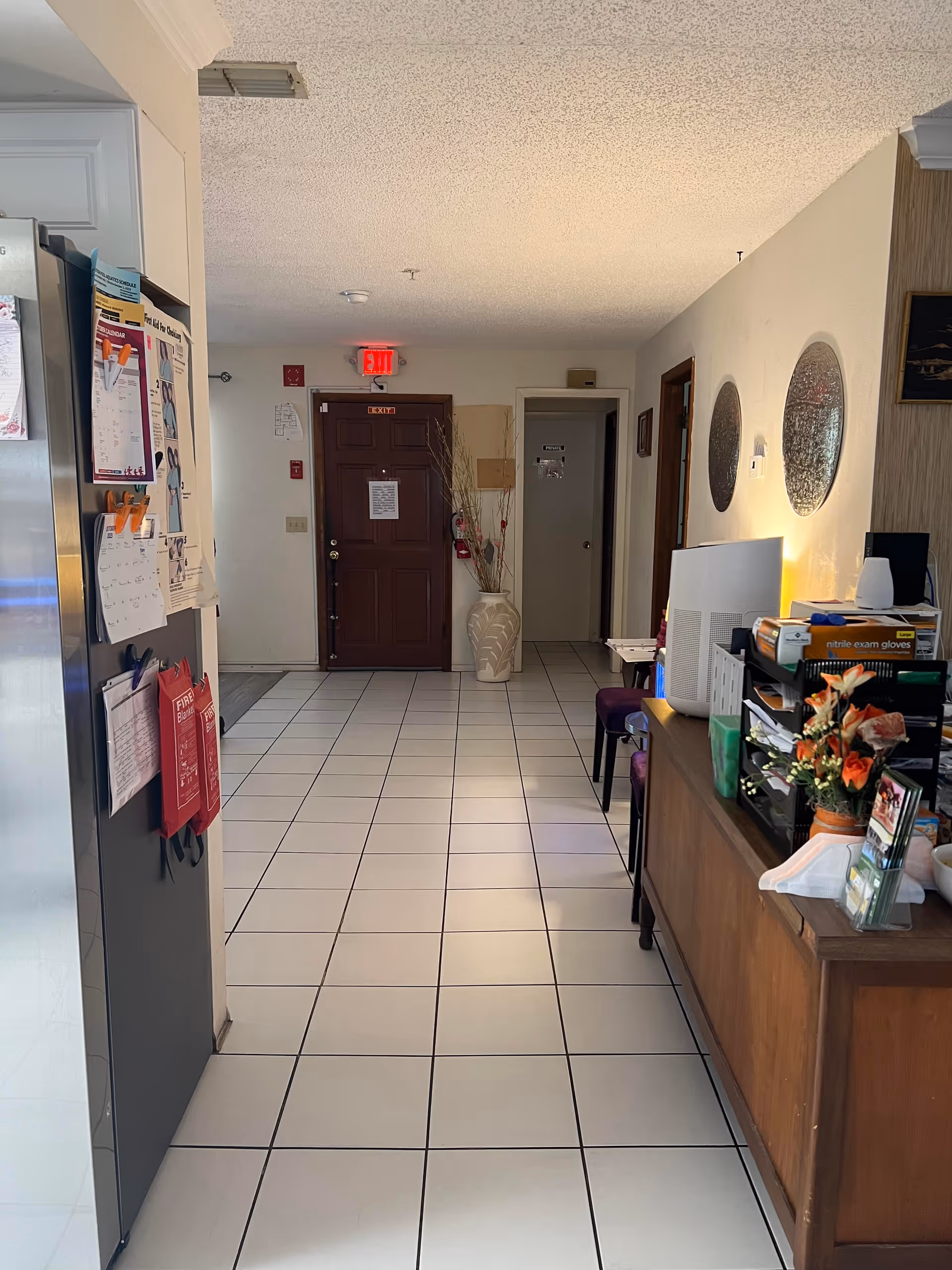 Interior hallway of a senior living facility with white tiled floor, a dark brown door at the end with an exit sign above it, a large decorative vase with dried branches next to the door, and a wooden reception desk on the right side with flowers, office supplies, and a computer monitor. On the left side, part of a refrigerator with papers and notes attached is visible.
