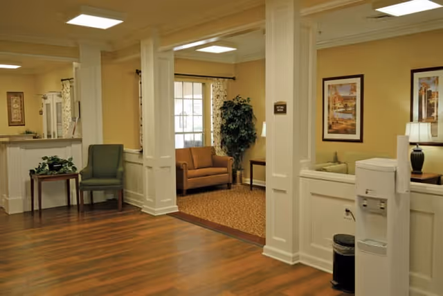Interior view of a senior living facility lounge area with wood flooring, beige walls, and white trim. The space includes a green upholstered chair, a brown loveseat, a side table with a plant, framed artwork on the walls, a water cooler, and a trash bin. Large windows with curtains allow natural light into the room.
