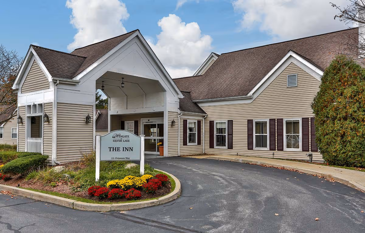 Exterior view of Wingate Residences at Silver Lake, showing the entrance to The Inn with a covered driveway, beige siding, brown roof, and a landscaped area with red and yellow flowers.
