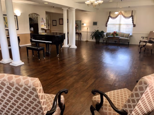 A spacious living room area with polished wooden floors, two patterned armchairs in the foreground, a black grand piano with a matching bench near white columns, and a seating area with a sofa and chairs near a window with blinds and valances. The room is softly lit with wall sconces and a ceiling light fixture.