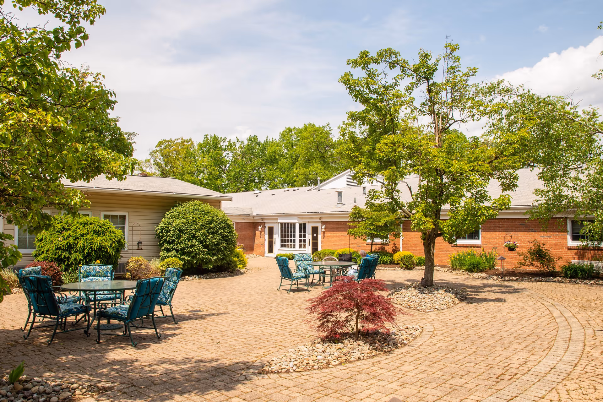 Outdoor courtyard area with paved brick ground, several green and blue cushioned metal chairs around glass tables, surrounded by trees and bushes, with a single-story building in the background under a partly cloudy sky.