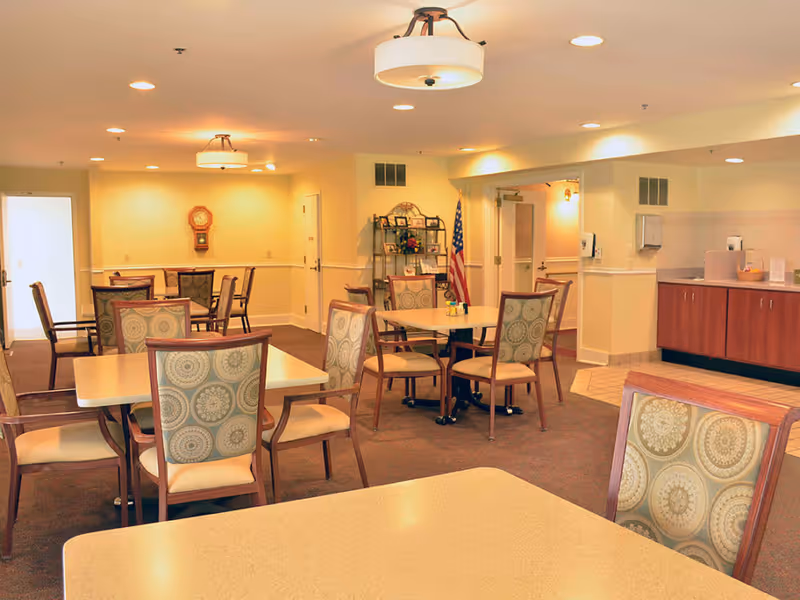A well-lit dining room with several tables and chairs arranged neatly. The chairs have patterned upholstery, and the room features warm beige walls, ceiling lights, a wall clock, an American flag, and a small shelf with decorative items. There is a counter area with cabinets on the right side.