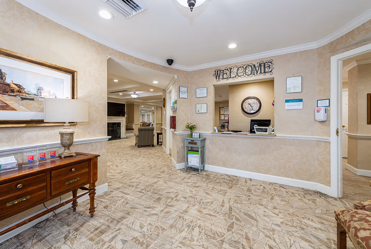 Reception area of a senior living facility with a welcome desk under a 'WELCOME' sign, seating visible in the background, patterned carpet and decorative furnishings.