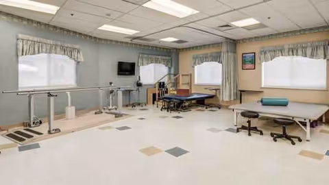 Bright rehabilitation therapy room with parallel bars, treatment tables, and exercise equipment.