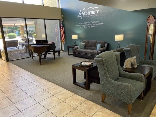 A senior living facility common area with a gray sofa, two patterned armchairs, side tables with lamps, a grand piano, an American flag, and a tall grandfather clock. The wall has the LifeStream logo and a mission statement. Large windows let in natural light.