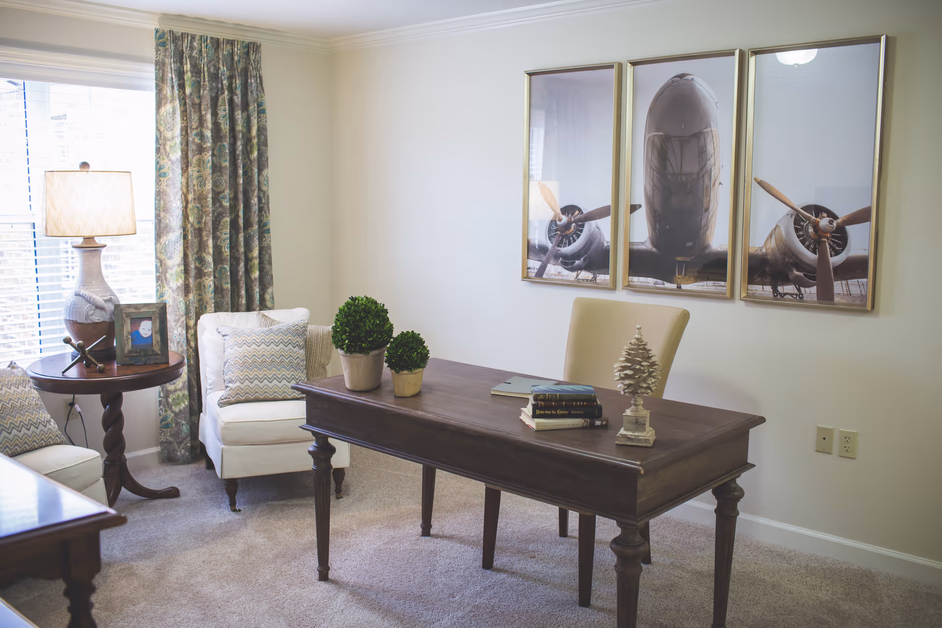 A light-filled sitting room with a wooden desk, upholstered chairs, a side table with a lamp, and a triptych airplane artwork on the wall.
