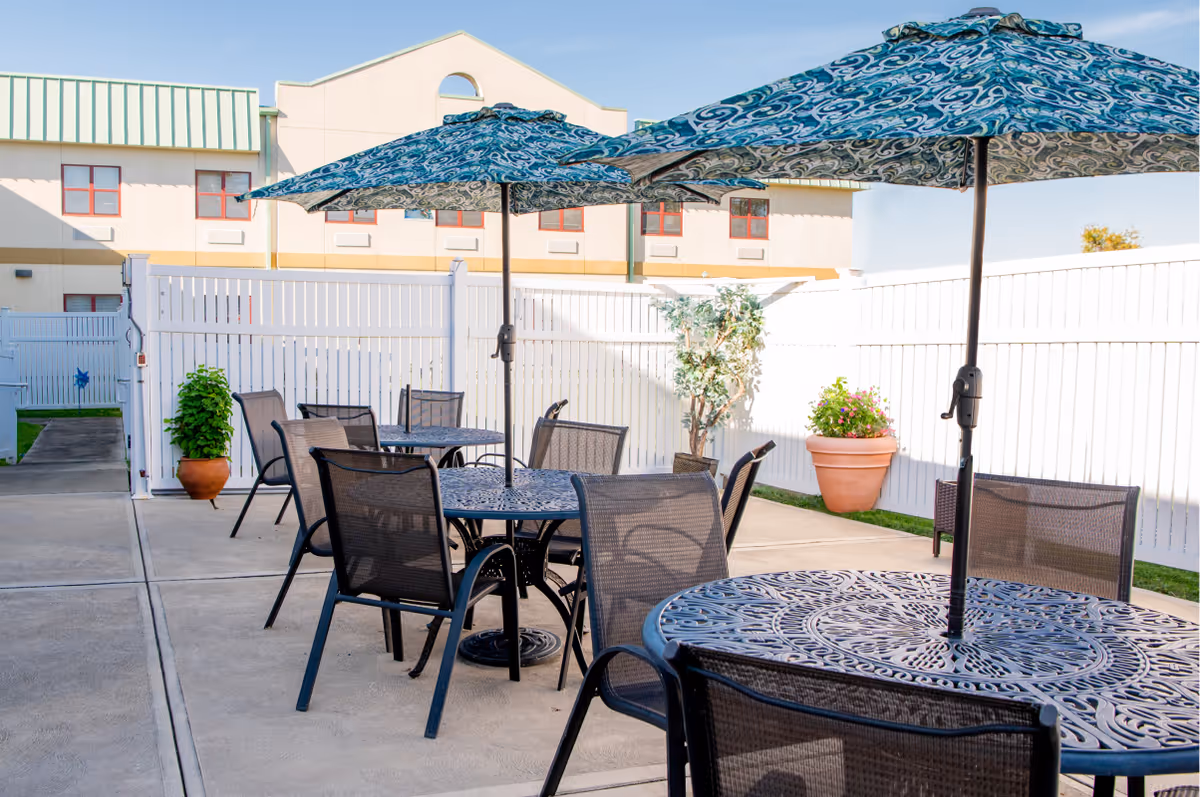 Outdoor patio area with several round metal tables and mesh chairs, each table shaded by large patterned umbrellas. The patio is enclosed by a white fence, with potted plants and a building visible in the background under a clear blue sky.