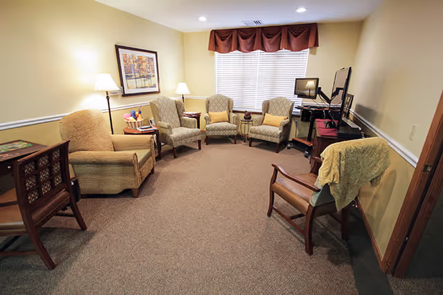 A cozy living room with six upholstered chairs arranged in a semi-circle around a television and computer setup. The room has beige walls, carpeted floor, two floor lamps, a framed picture on the wall, and a window with blinds and a valance.