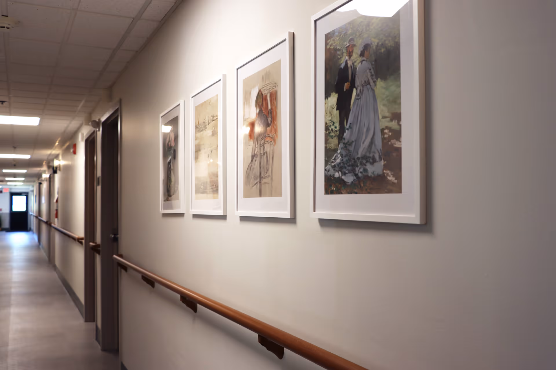 A well-lit hallway in a senior living facility with handrails on both sides and framed artwork hanging on the right wall. Several doors line the left side of the hallway, and the floor is covered with smooth, light-colored tiles.