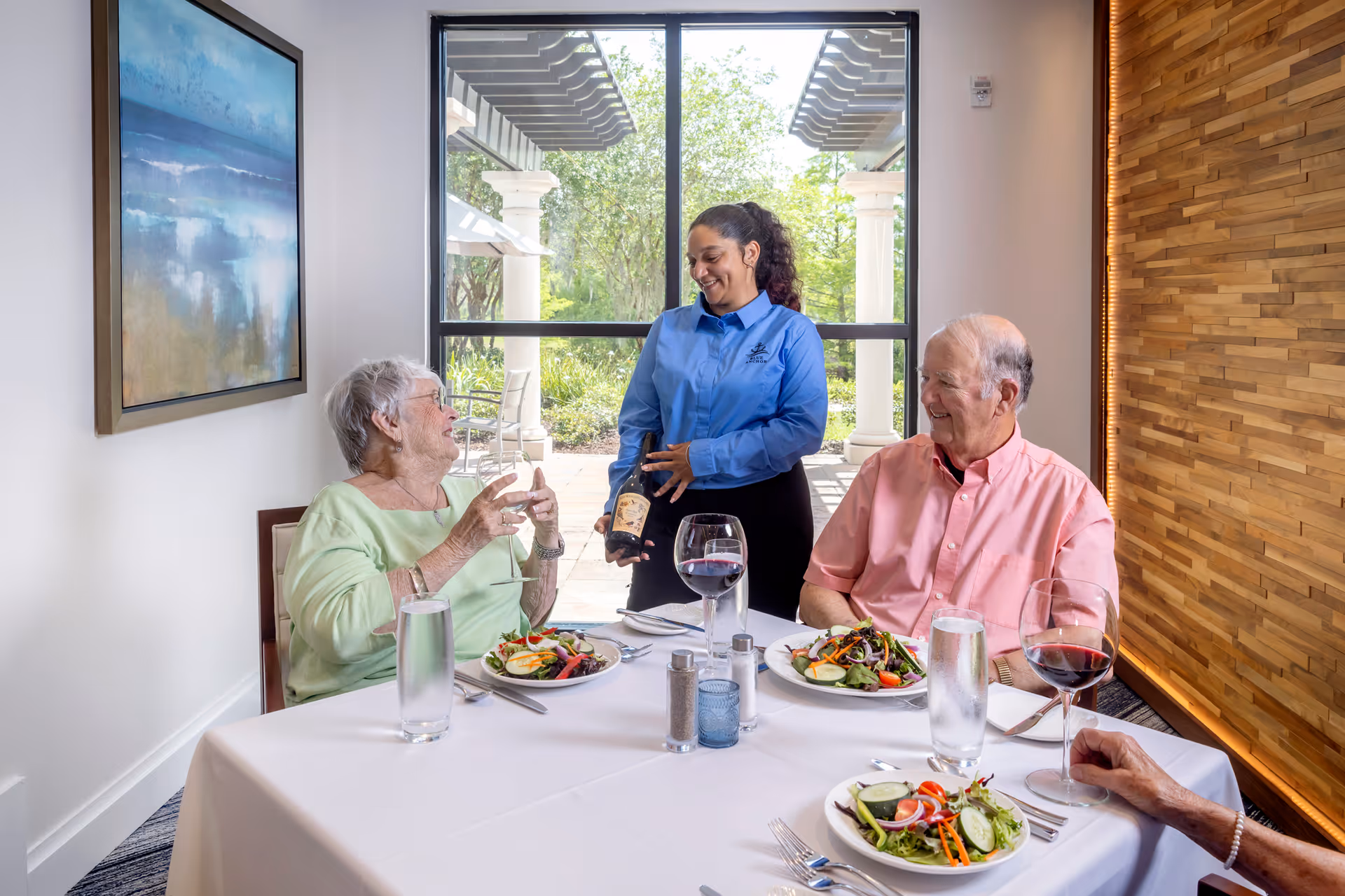A server pours wine for two elderly residents seated at a dining table with salads in a bright dining room with a large window and wood accent wall.