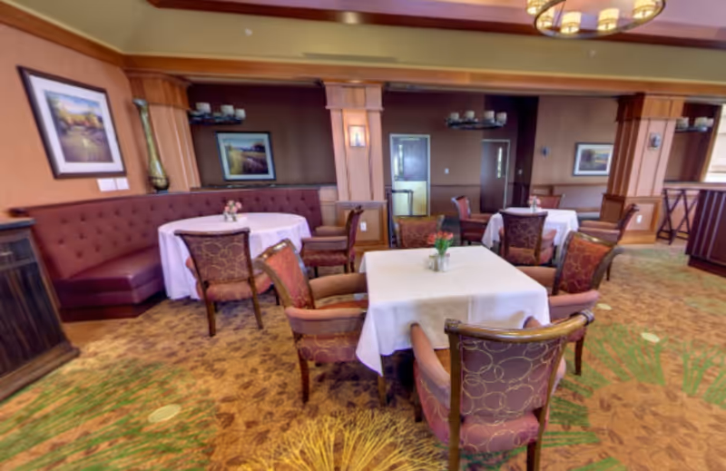 A cozy dining room with multiple tables covered in white tablecloths, surrounded by upholstered chairs with patterned fabric. The room features warm wood paneling, framed landscape artwork on the walls, and soft lighting from ceiling fixtures. There is a built-in bench seat along one wall and decorative plants on the carpeted floor.